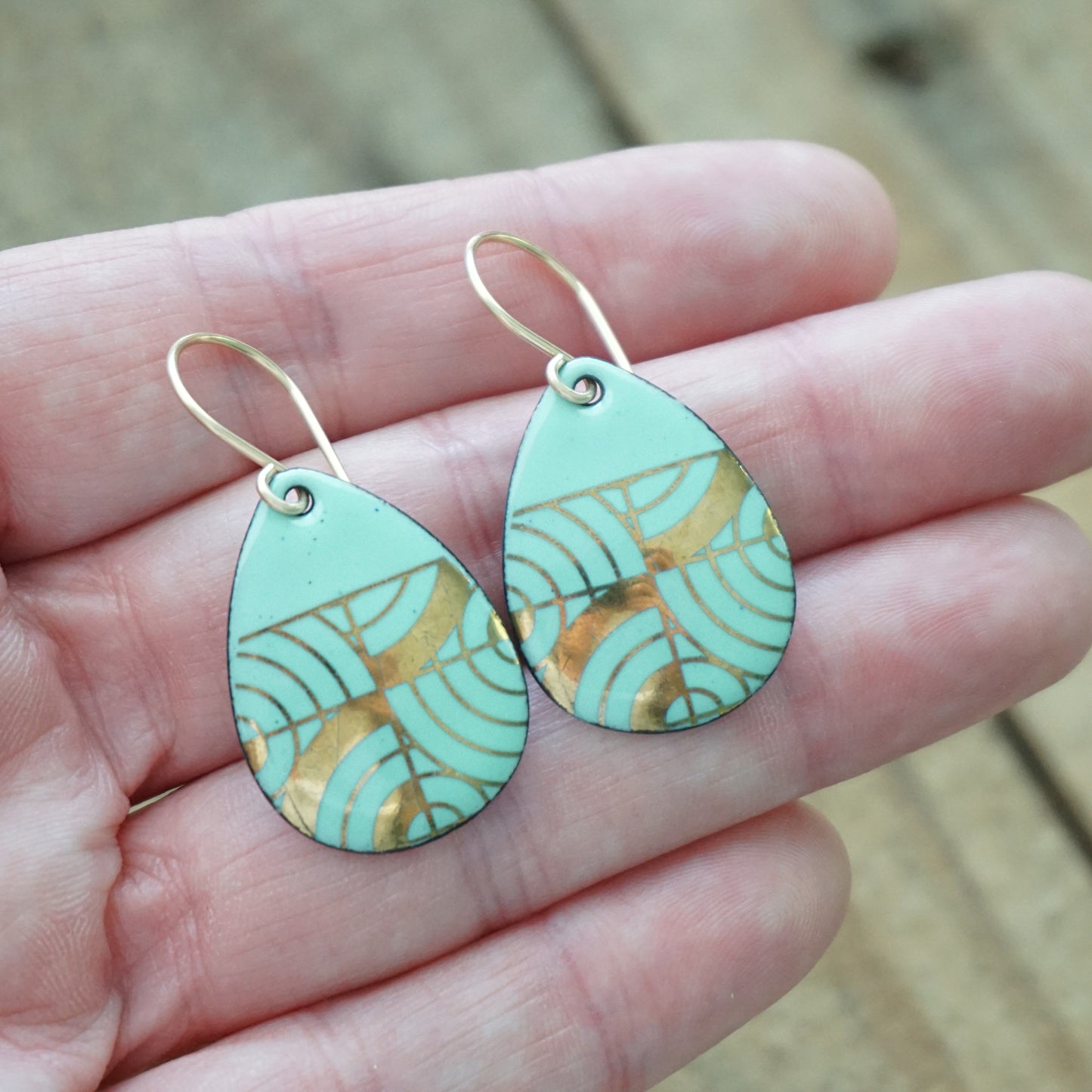Hand holding A pair of teardrop-shaped dangle earrings with seafoam green enamel and gold geometric pattern, against a woodent background
