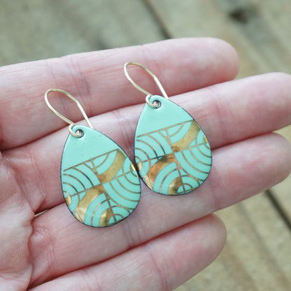 Hand holding A pair of teardrop-shaped dangle earrings with seafoam green enamel and gold geometric pattern, against a woodent background