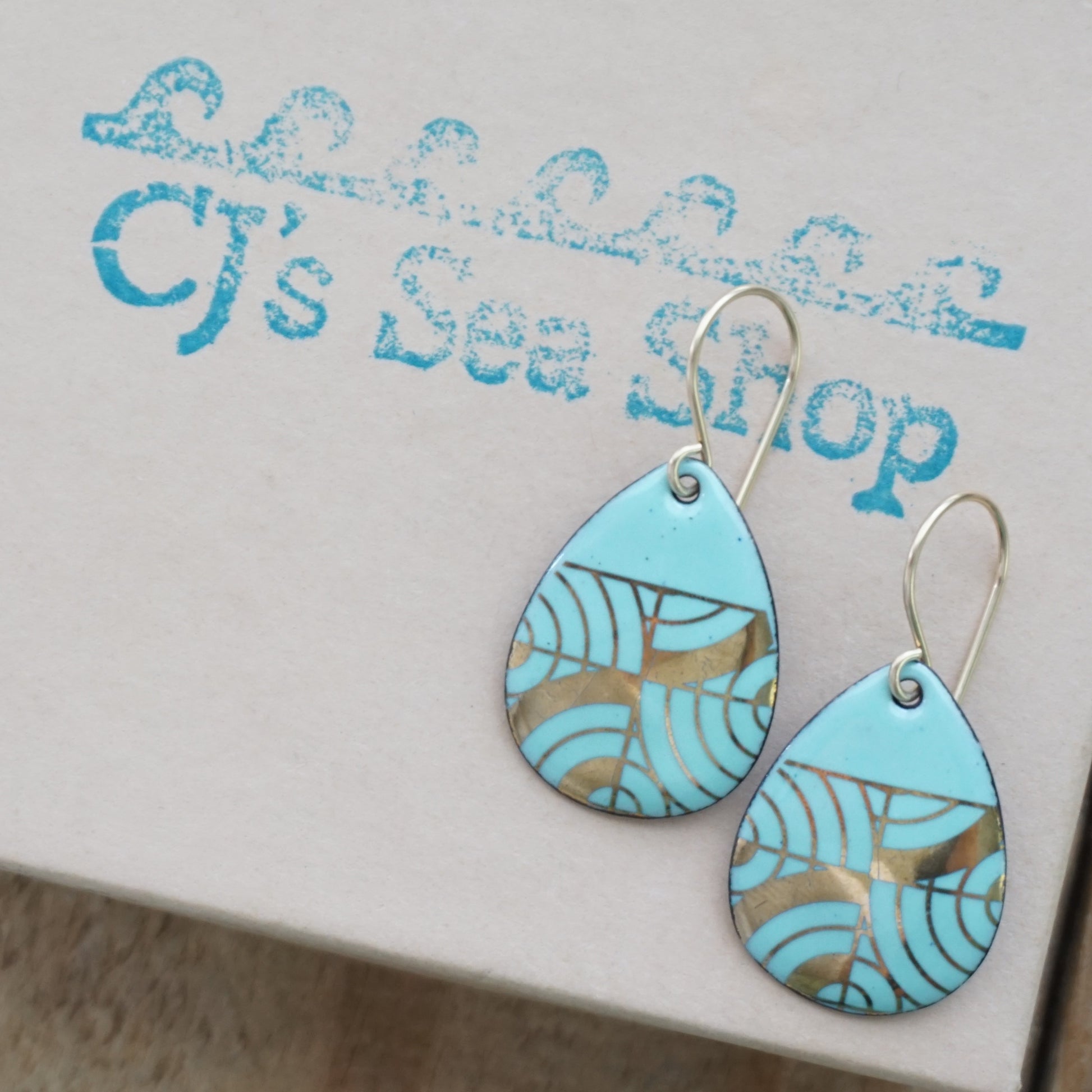 A pair of teardrop-shaped dangle earrings with seafoam green enamel and gold geometric pattern, displayed on a gift box