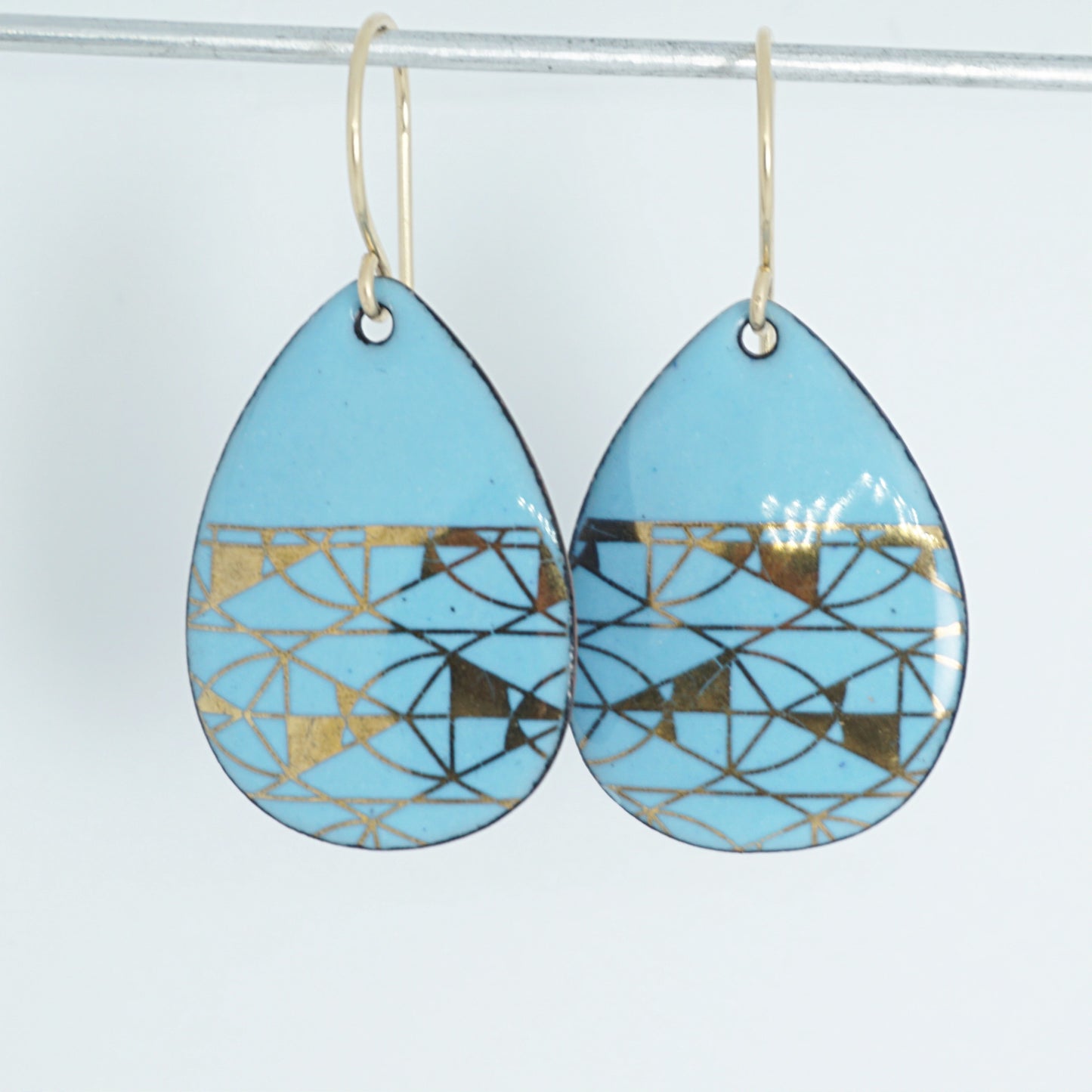 A pair of teardrop-shaped dangle earrings with a light blue enamel background, featuring a metallic geometric pattern, hanging from thin gold hooks.