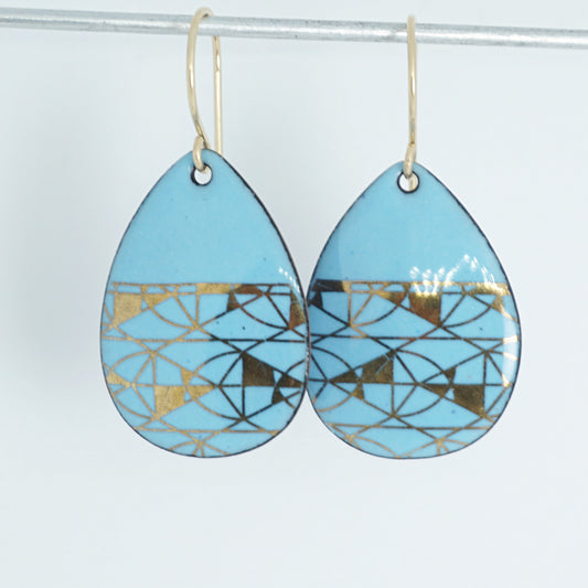 A pair of teardrop-shaped dangle earrings with a light blue enamel background, featuring a metallic geometric pattern, hanging from thin gold hooks.