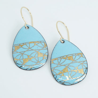 A pair of teardrop-shaped dangle earrings with a light blue enamel background, featuring a metallic geometric pattern, hanging from thin gold hooks.