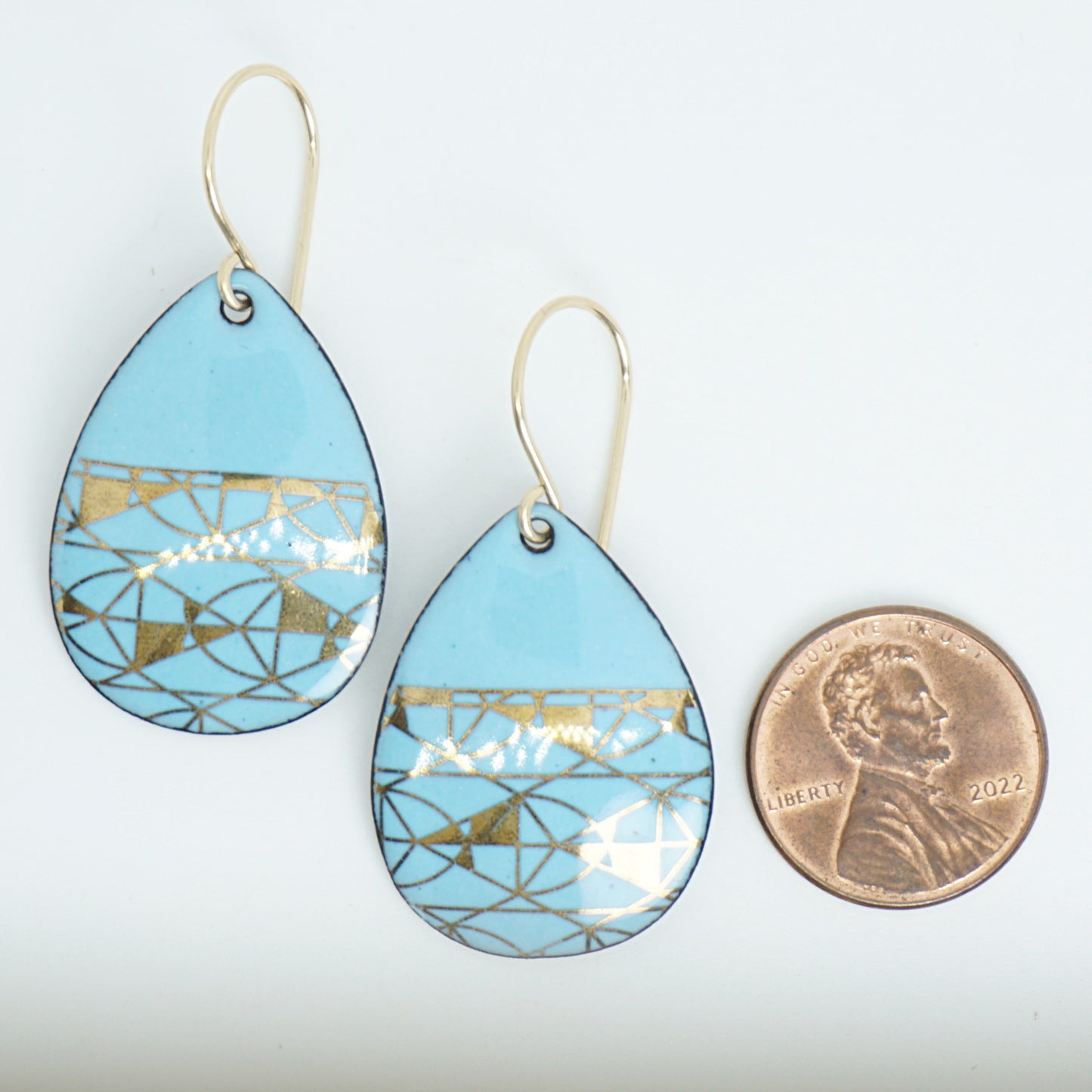 A pair of teardrop-shaped dangle earrings with a light blue enamel background, featuring a metallic geometric pattern, hanging from thin gold hooks, next to a penny for scale, against a white background