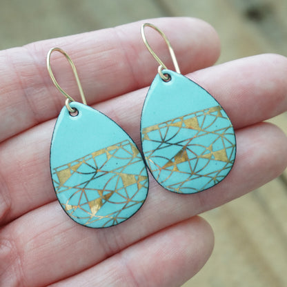 Hand holding A pair of teardrop-shaped dangle earrings with a light blue enamel background, featuring a metallic geometric pattern, hanging from thin gold hooks, against a woodent background