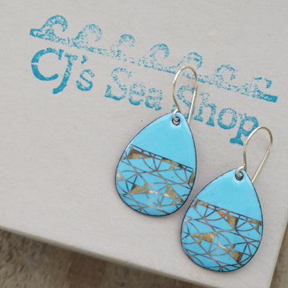 A pair of teardrop-shaped dangle earrings with a light blue enamel background, featuring a metallic geometric pattern, hanging from thin gold hooks, displayed on a gift box