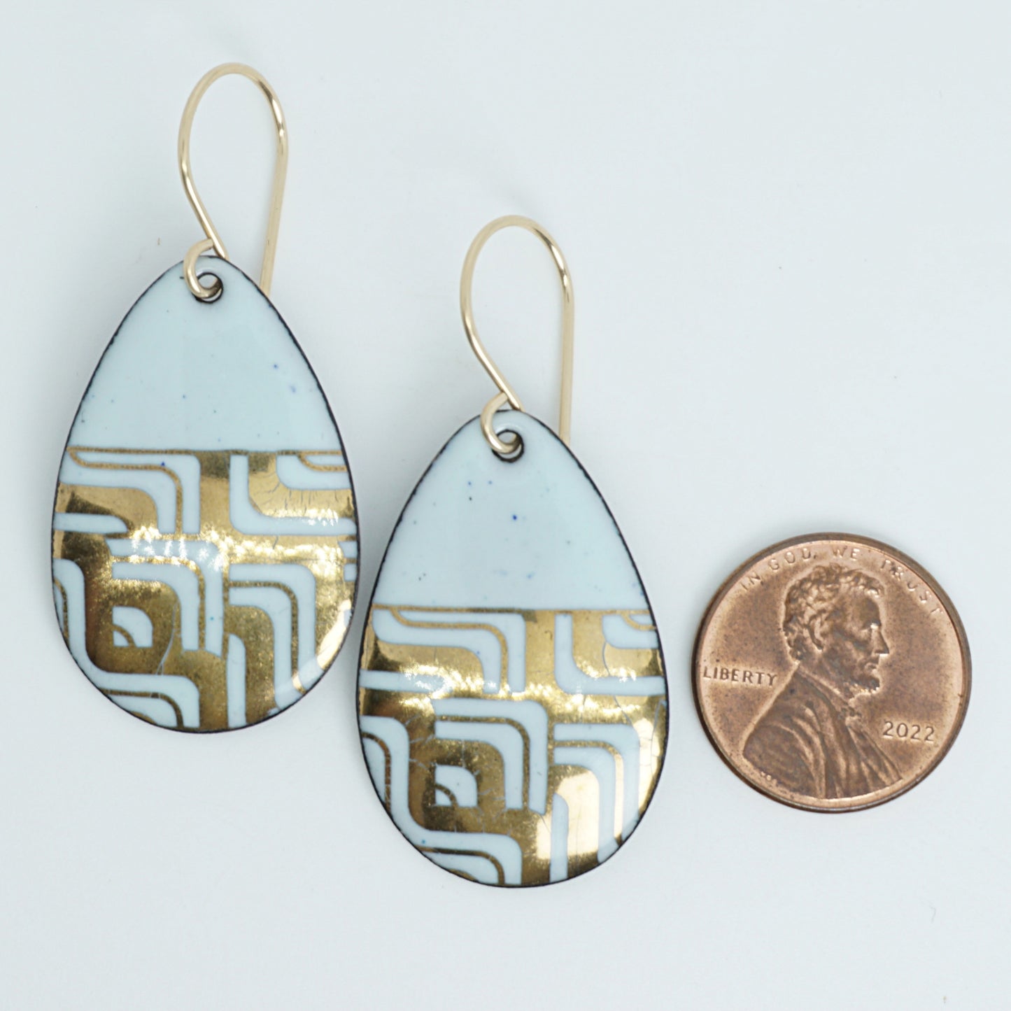 A pair of teardrop-shaped dangle earrings with seafoam green enamel and gold geometric pattern, next to a penny for scale, against a white background