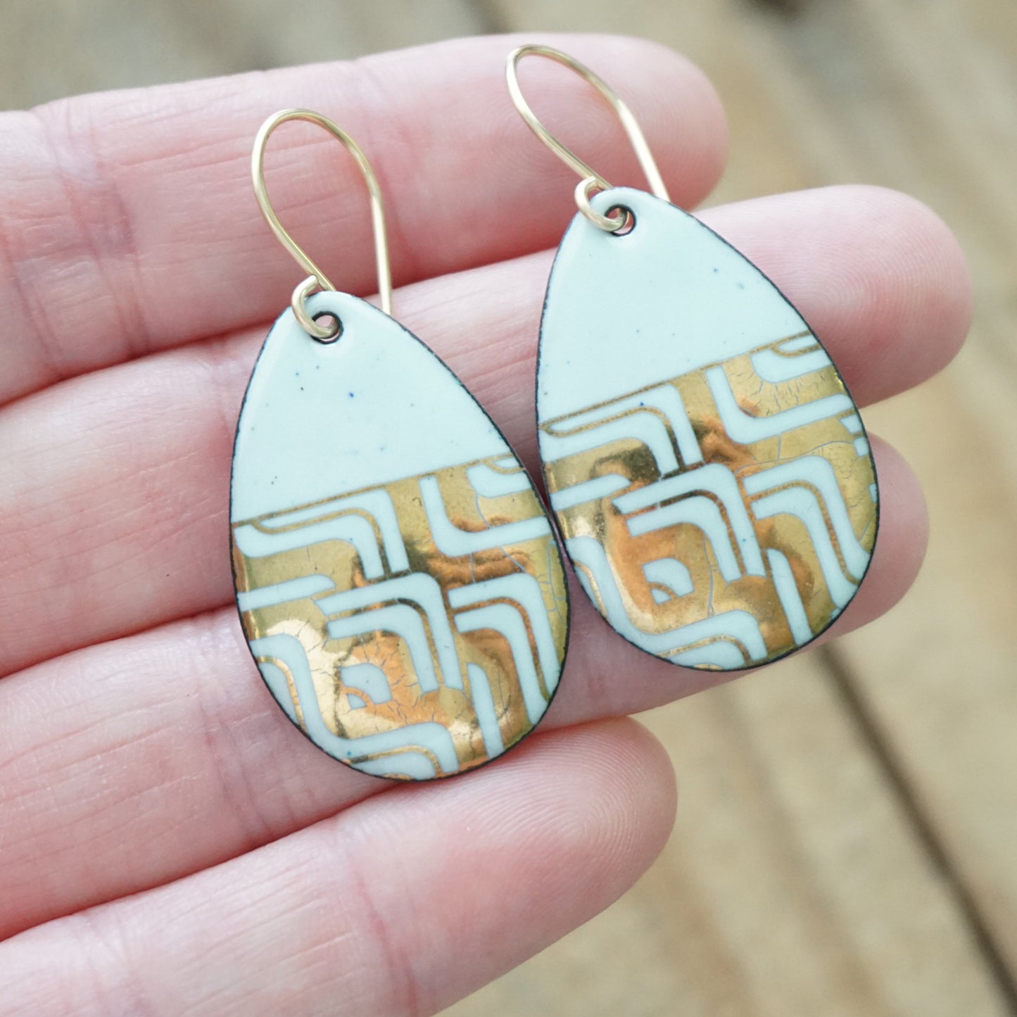 hand holding A pair of teardrop-shaped dangle earrings with seafoam green enamel and gold geometric pattern, against a wooden background