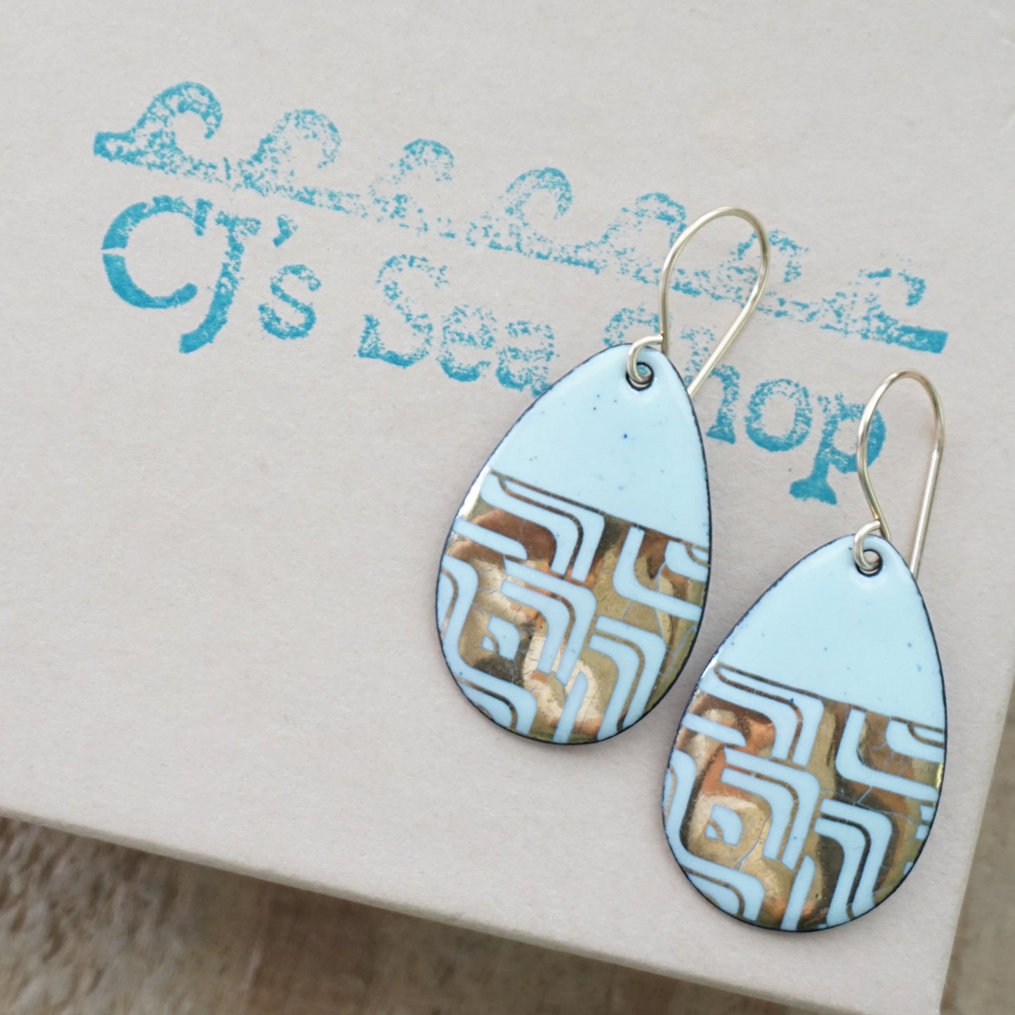 A pair of teardrop-shaped dangle earrings with seafoam green enamel and gold geometric pattern, displayed on a gift box
