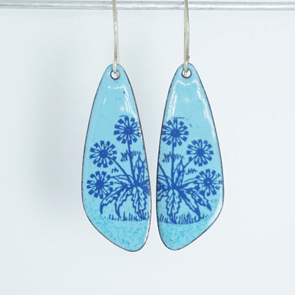 A pair of light blue teardrop earrings with blue floral dandelion designs, against a white background