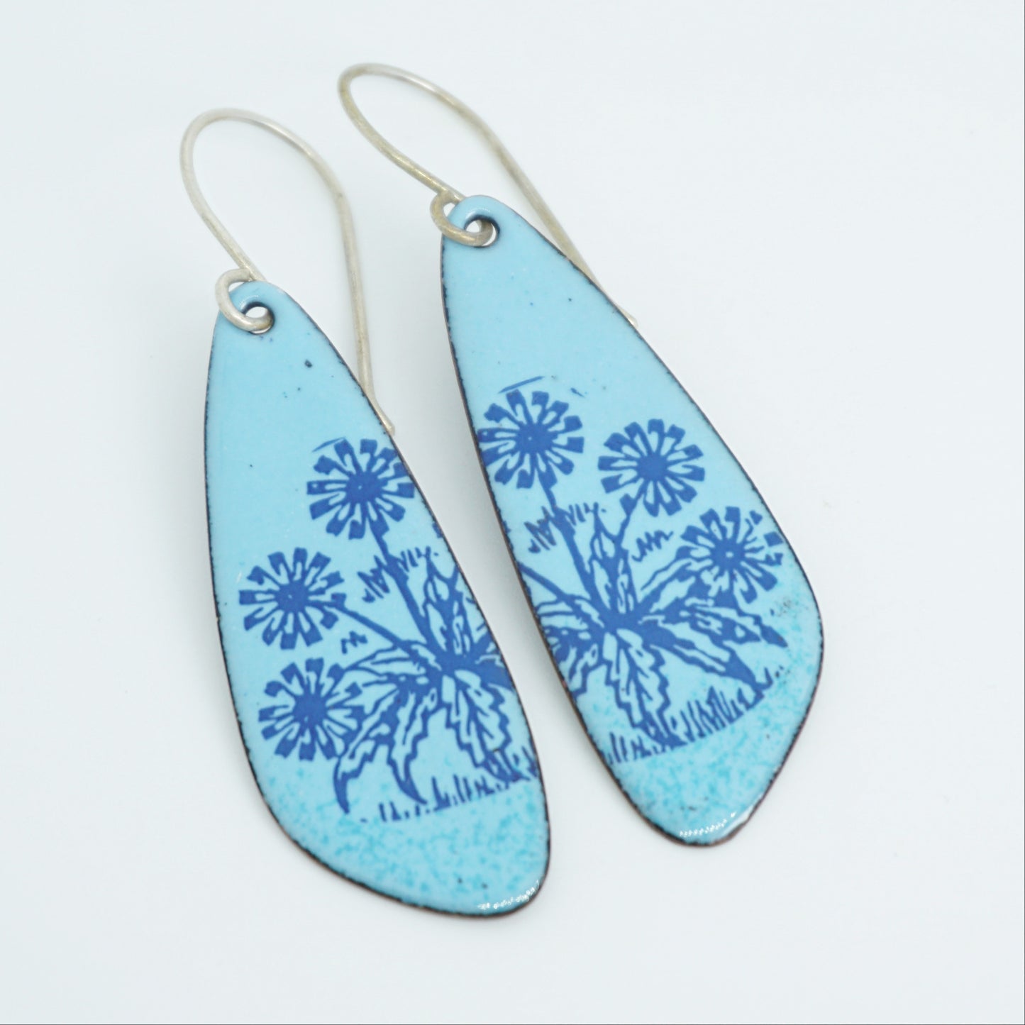 A pair of light blue teardrop earrings with blue floral dandelion designs, against a white background