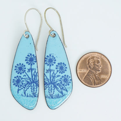 A pair of light blue teardrop earrings with blue floral dandelion designs, next to a penny for scale, against a white background