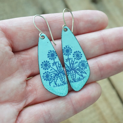 Hand holding A pair of light blue teardrop earrings with blue floral dandelion designs, against a wooden background