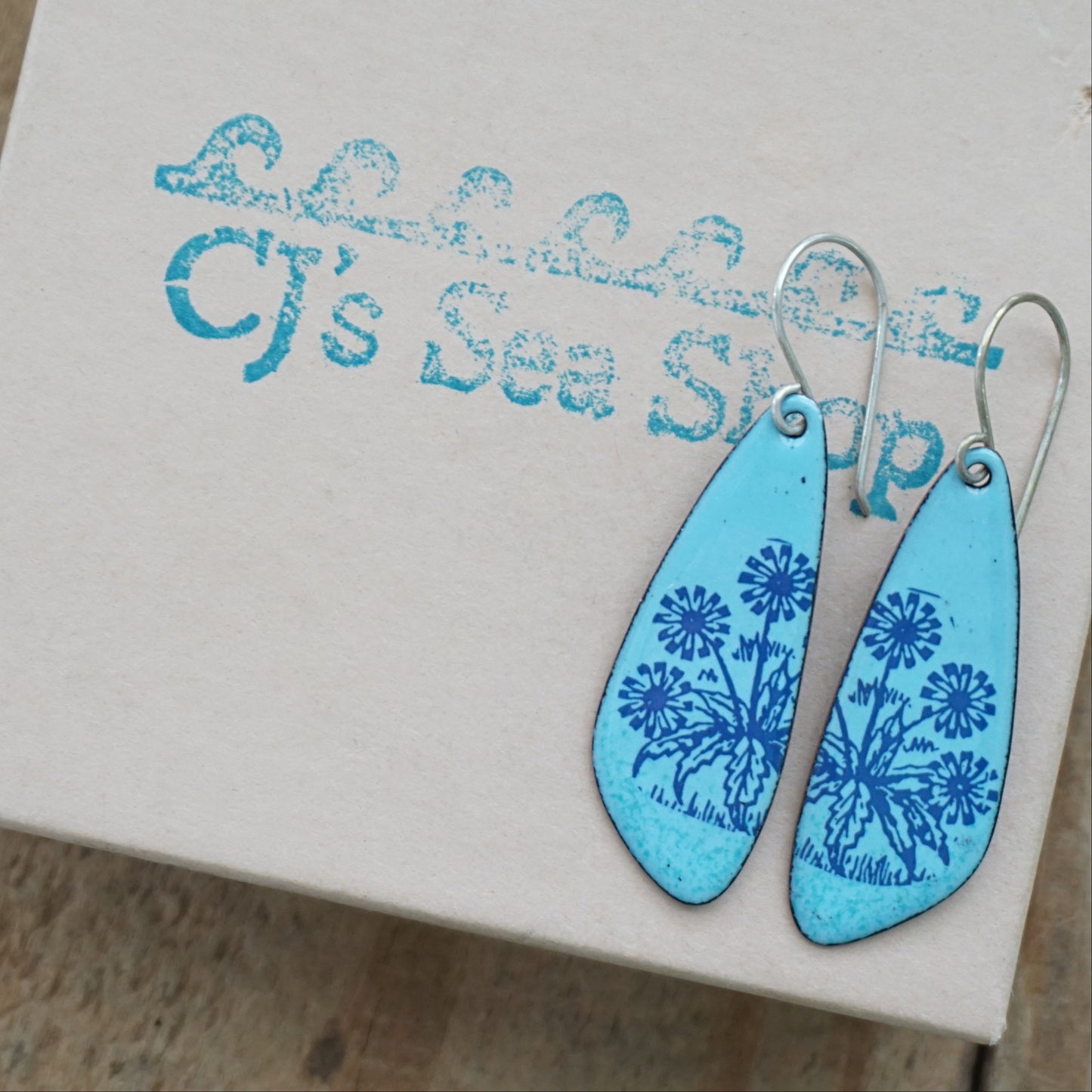 A pair of light blue teardrop earrings with blue floral dandelion designs, displayed on a gift box