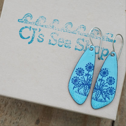 A pair of light blue teardrop earrings with blue floral dandelion designs, displayed on a gift box