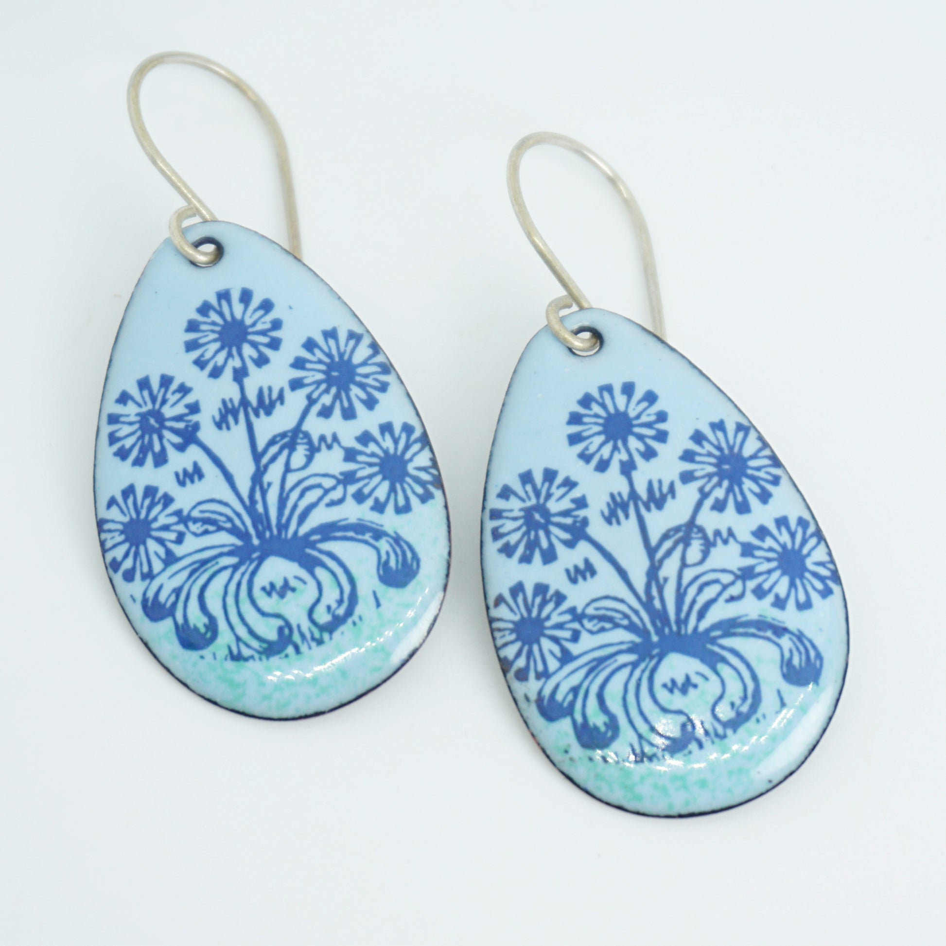 A pair of light blue teardrop earrings with blue floral dandelion designs, against a white background