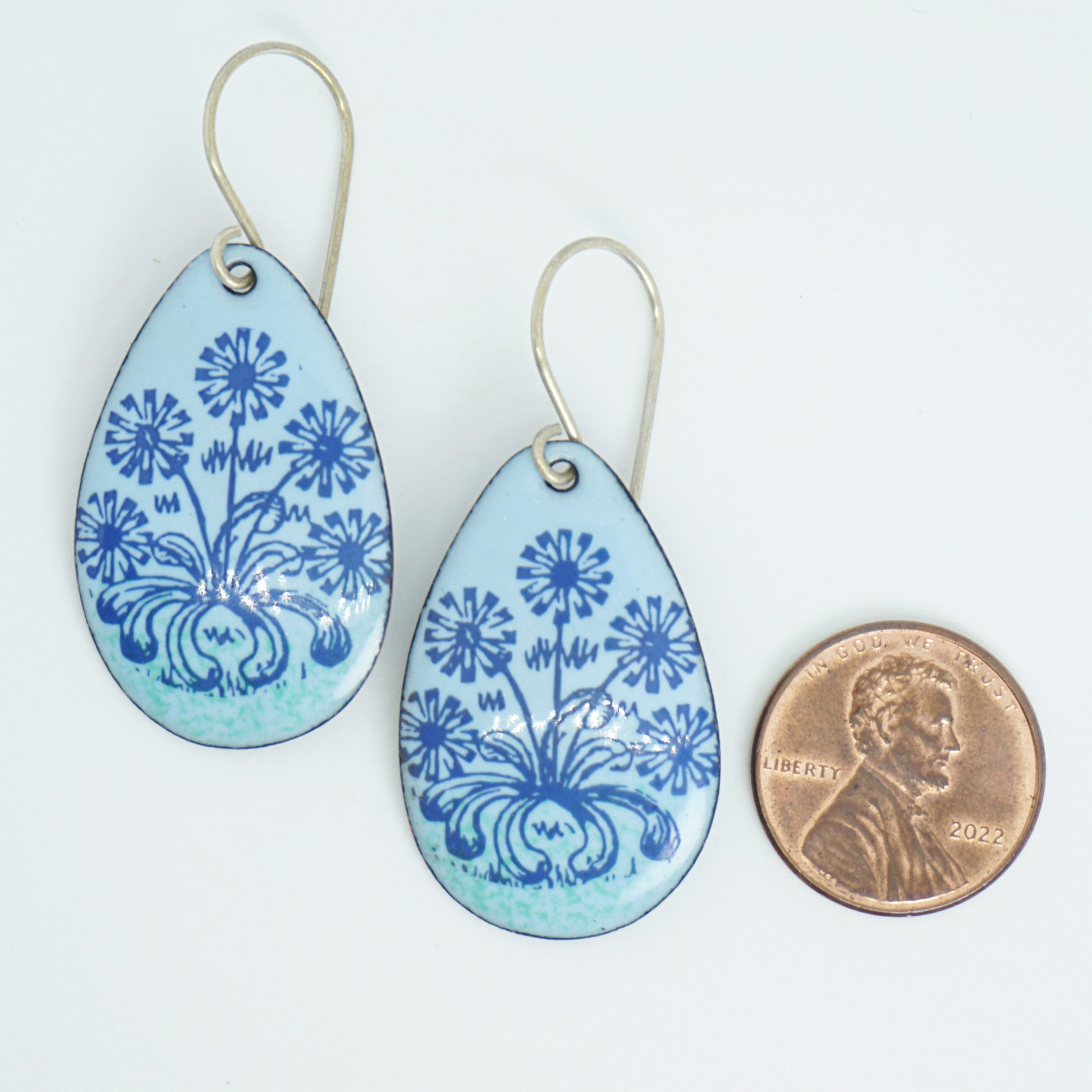 A pair of light blue teardrop earrings with blue floral dandelion designs, next to a penny for scale, against a white background