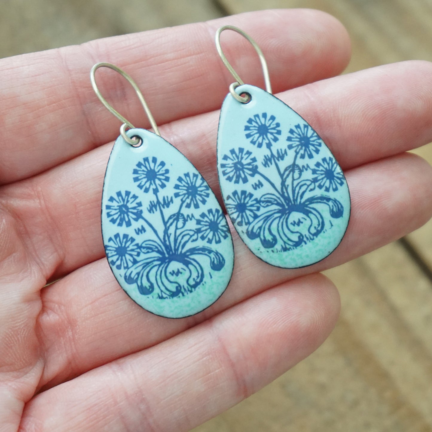 Hand holding A pair of light blue teardrop earrings with blue floral dandelion designs, against a wooden background