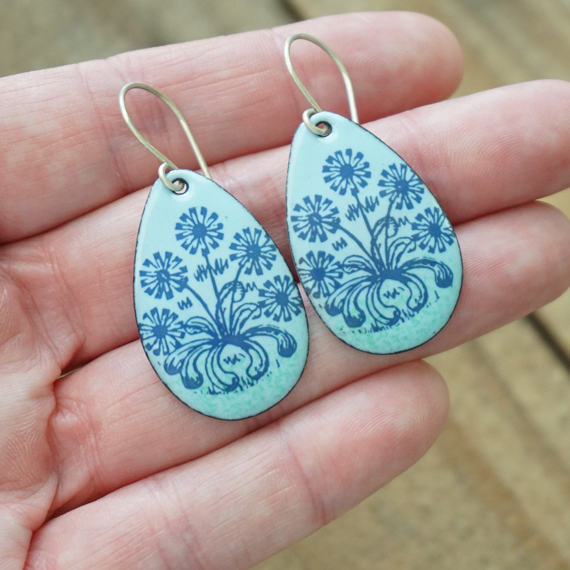 Hand holding A pair of light blue teardrop earrings with blue floral dandelion designs, against a wooden background