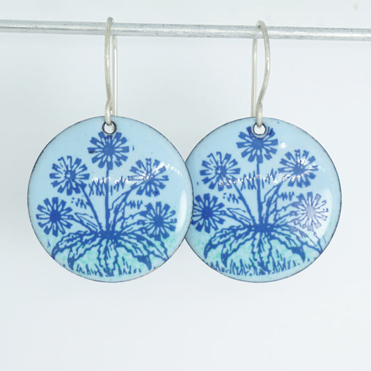 A pair of light blue round earrings with blue floral dandelion designs, against a white background.