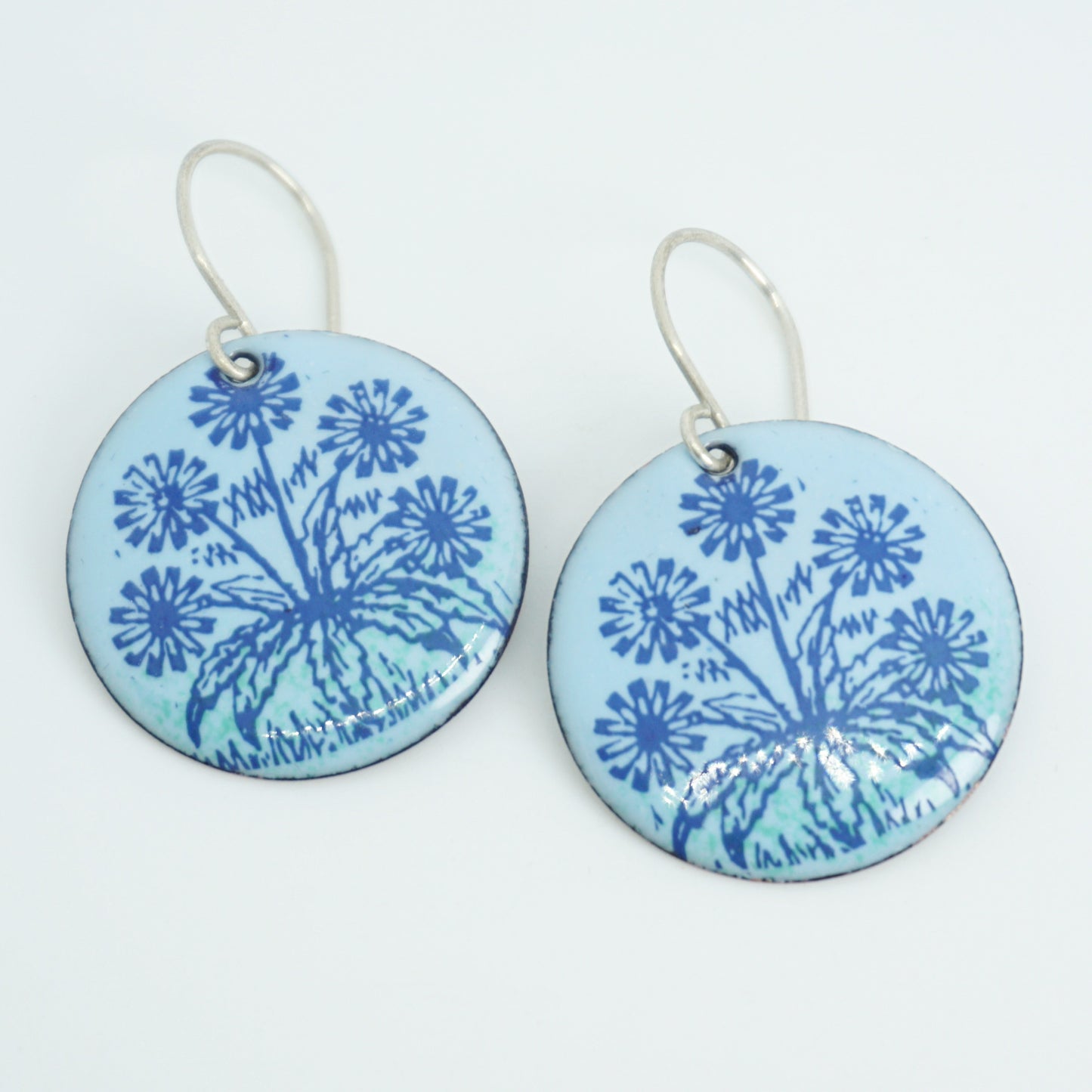 A pair of light blue round earrings with blue floral dandelion designs, against a white background.