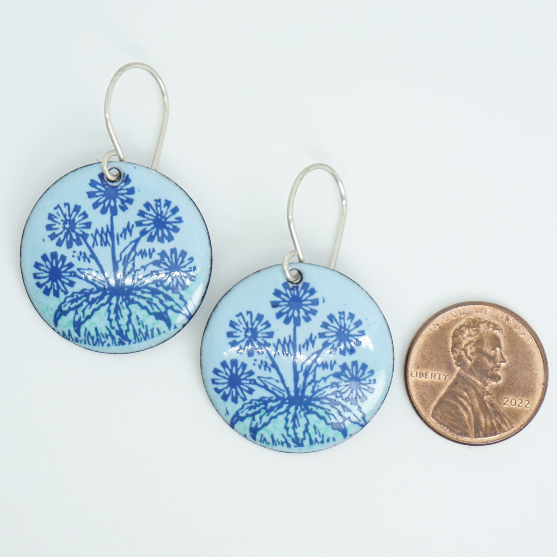 A pair of light blue round earrings with blue floral dandelion designs, next to a penny for scale, against a white background.