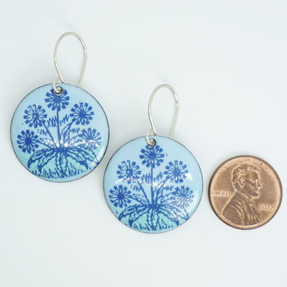 A pair of light blue round earrings with blue floral dandelion designs, next to a penny for scale, against a white background.