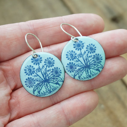 hand holding A pair of light blue round earrings with blue floral dandelion designs, against a wooden background.