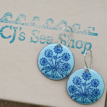 A pair of light blue round earrings with blue floral dandelion designs, displayed on a gift box