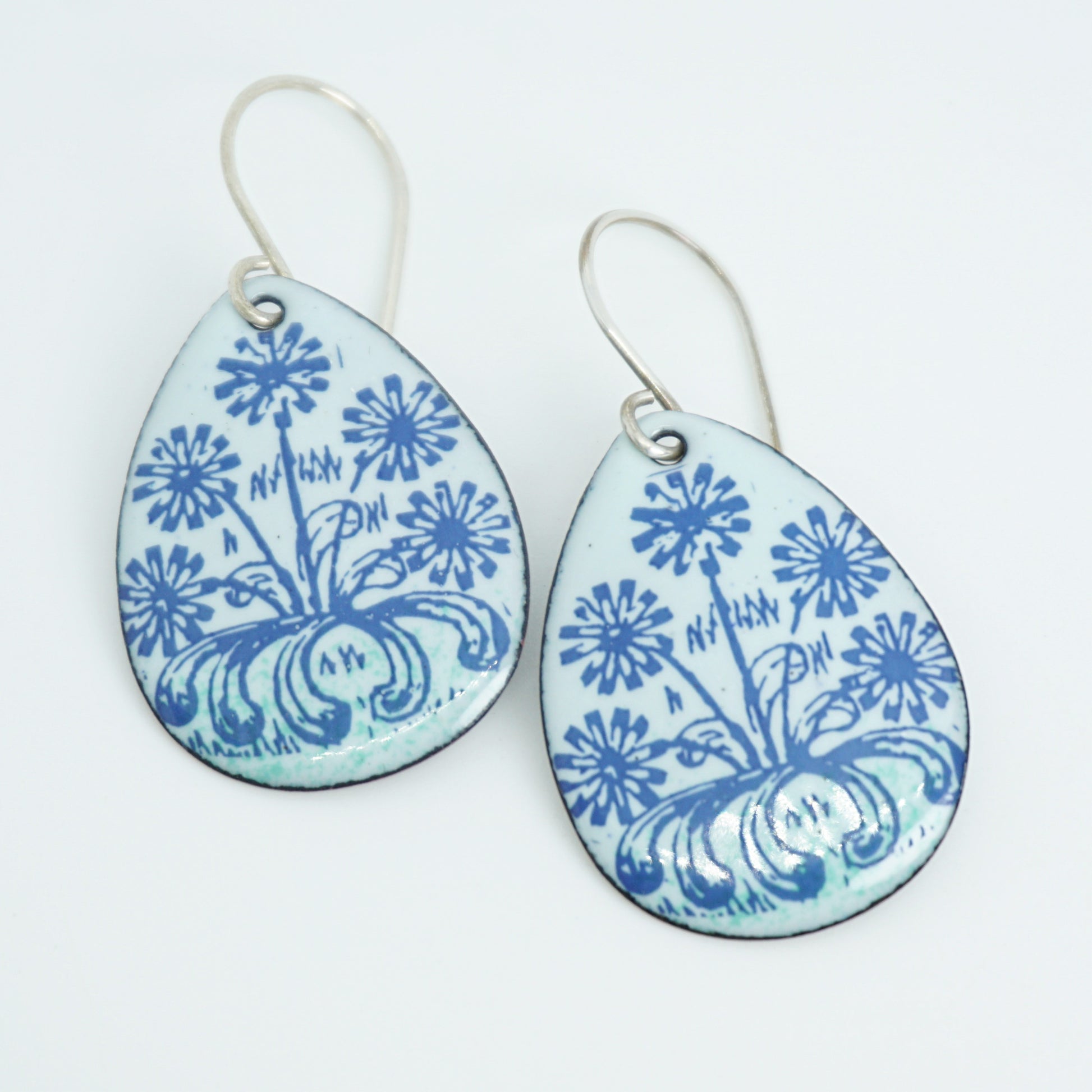 A pair of light seafoam green teardrop earrings with blue floral dandelion designs, against a white background