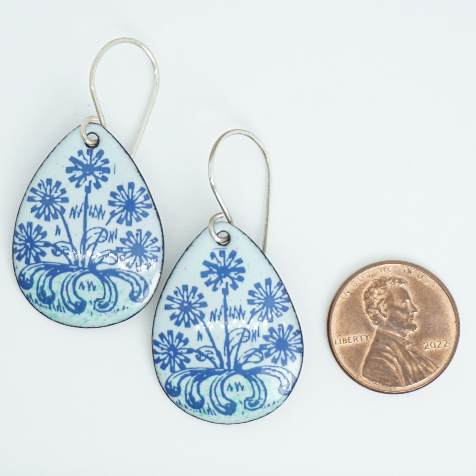 A pair of light seafoam green teardrop earrings with blue floral dandelion designs, next to a penny for scale, against a white background