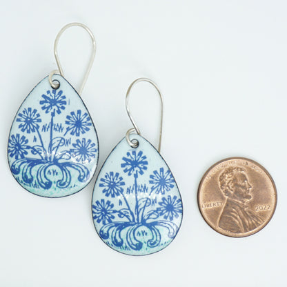 A pair of light seafoam green teardrop earrings with blue floral dandelion designs, next to a penny for scale, against a white background