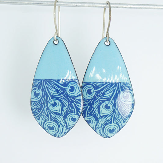 A pair of teardrop-shaped drop earrings with a light blue top half and a blue peacock-feather pattern bottom half, hanging from silver hooks against a plain white background.