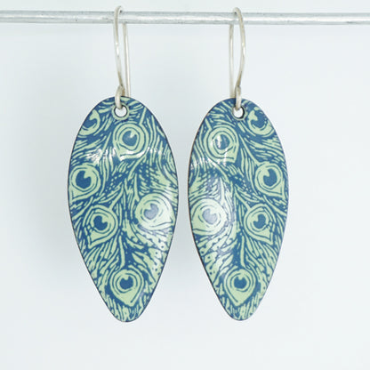 A pair of teardrop-shaped dangling earrings with a blue and light green peacock feather pattern, hanging from silver hooks against a plain white background.