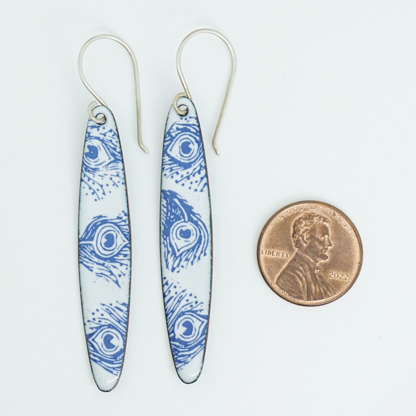 A pair of long white dangle earrings with blue peacock feather patterns and silver fishhook wires, shown next to a US penny for scale on a white background.