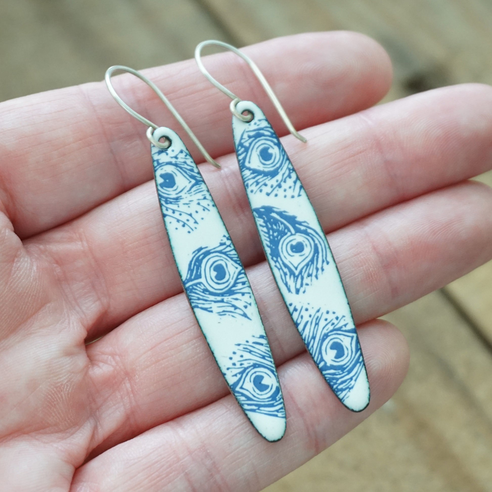 A pair of long, narrow dangle earrings with a blue peacock feather pattern on a white background, hanging from silver fishhook wires and held in a person's hand.