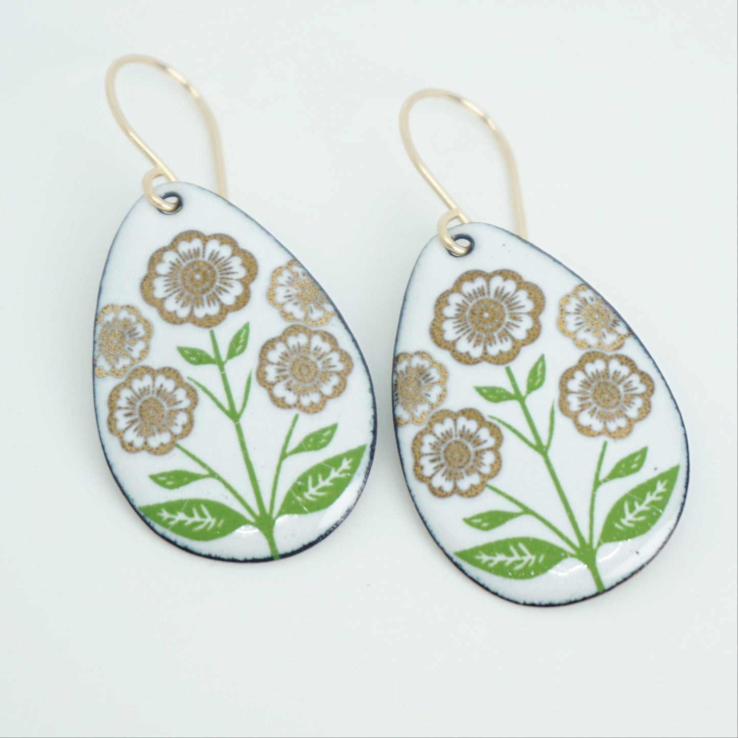 White teardrop-shaped dangle earrings, with gold flowers on green stems dangling from gold hooks, against a white background
