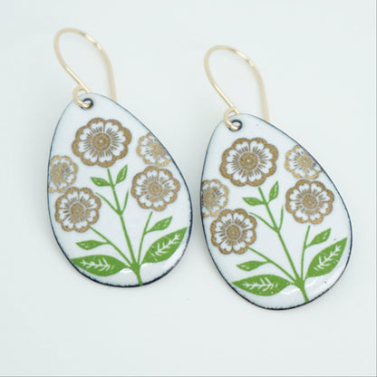 White teardrop-shaped dangle earrings, with gold flowers on green stems dangling from gold hooks, against a white background