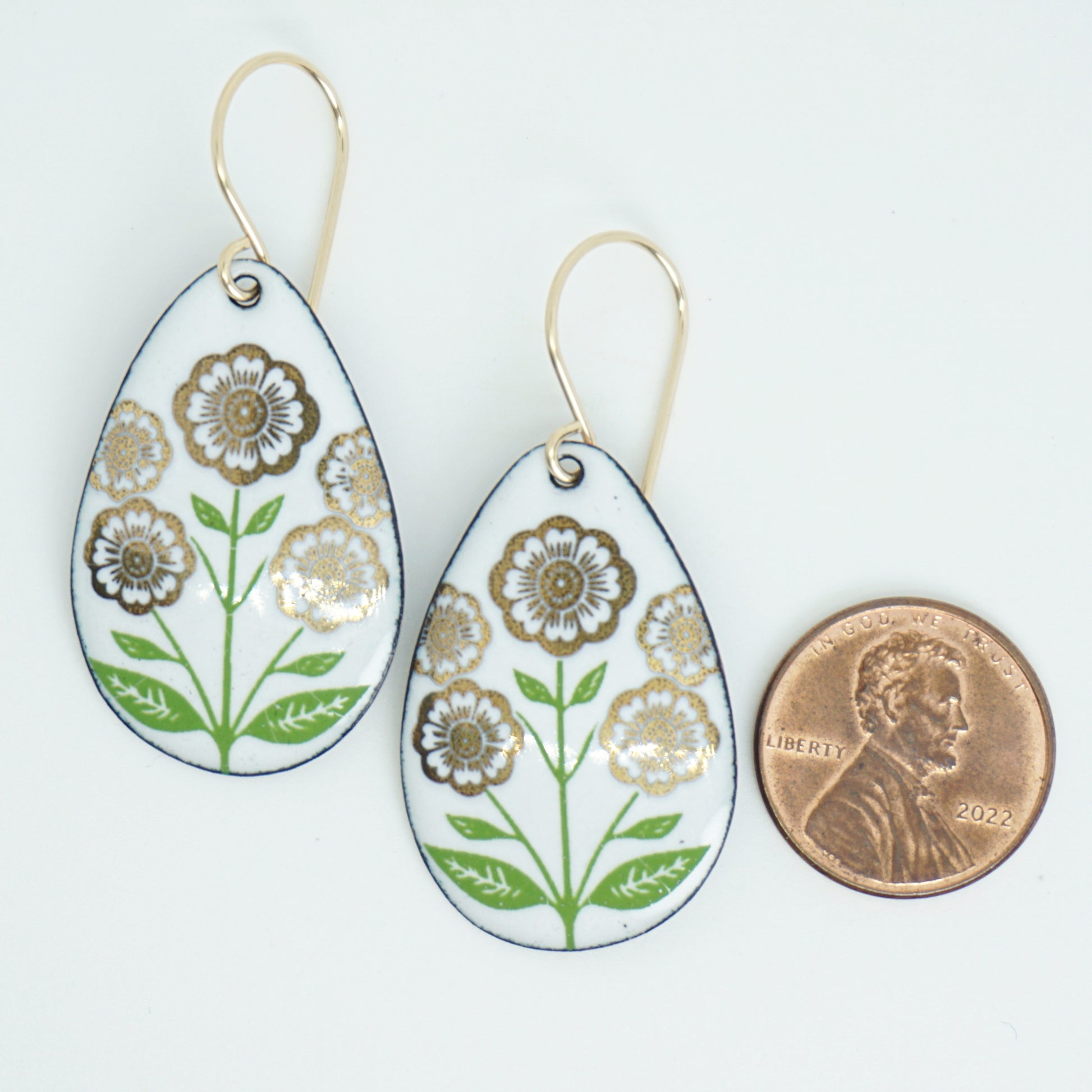 White teardrop-shaped dangle earrings, with gold flowers on green stems dangling from gold hooks, next to a penny for scale, against a white background