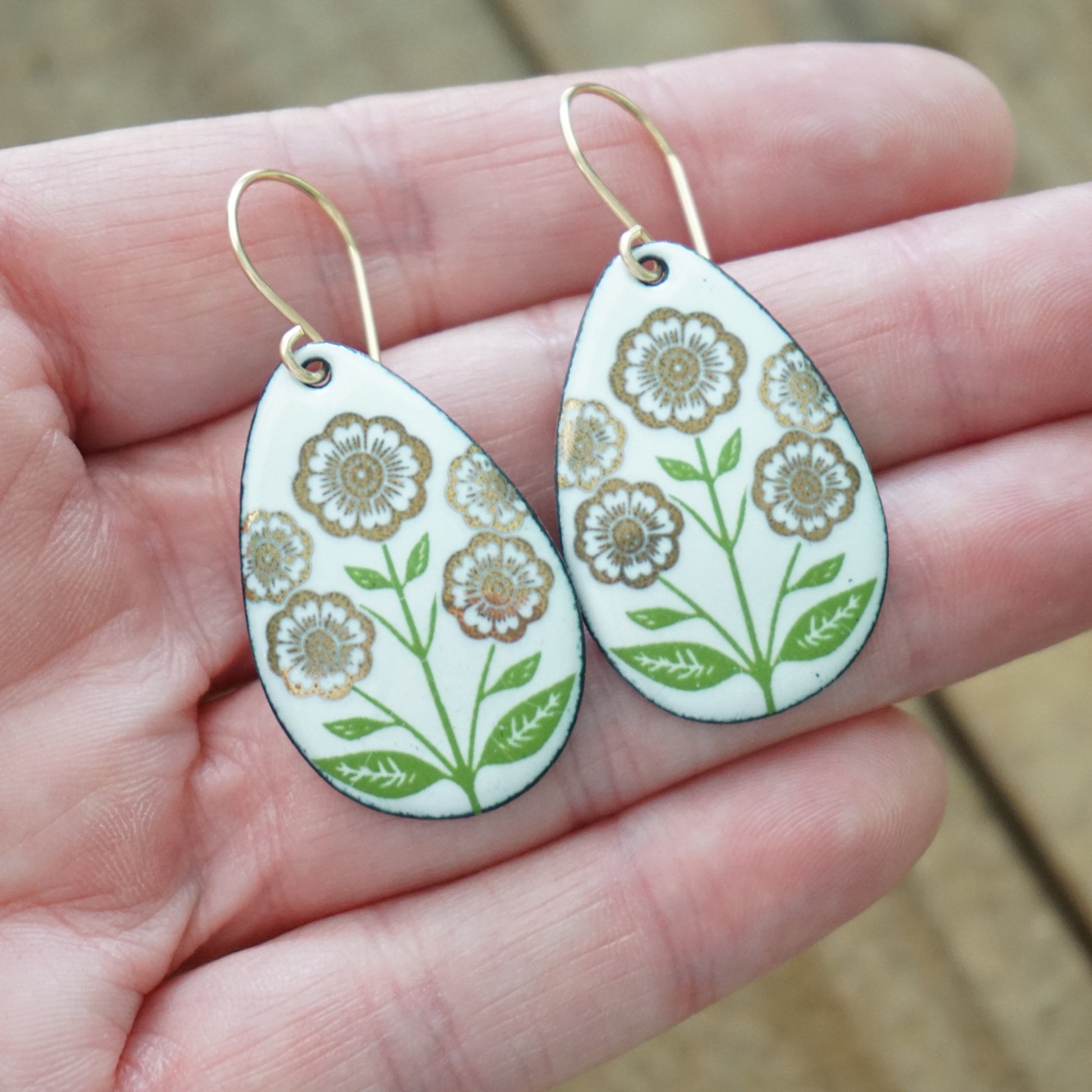 Hand holding a pair of White teardrop-shaped dangle earrings, with gold flowers on green stems dangling from gold hooks, against a wooden background