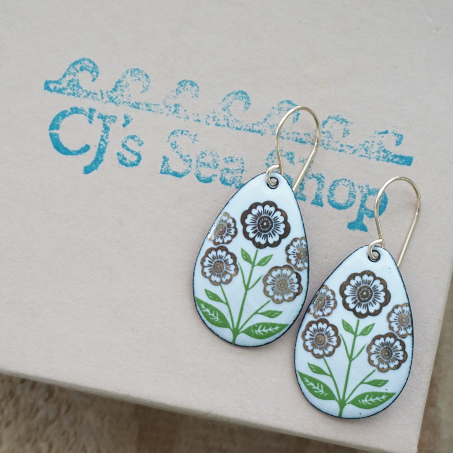 White teardrop-shaped dangle earrings, with gold flowers on green stems dangling from gold hooks, displayed on a gift box