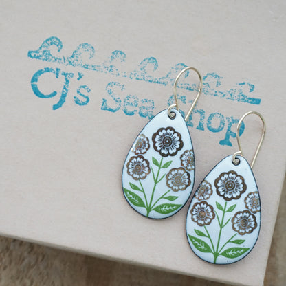 White teardrop-shaped dangle earrings, with gold flowers on green stems dangling from gold hooks, displayed on a gift box