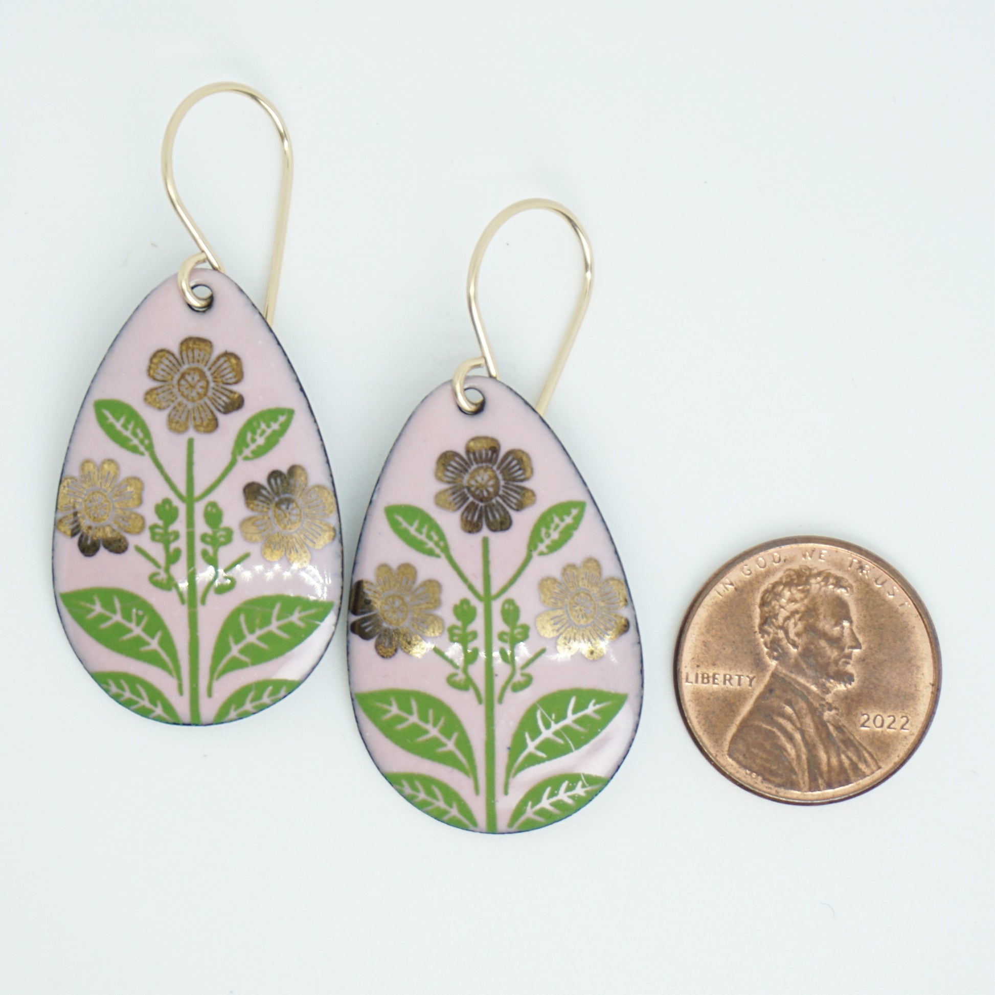 Pink teardrop-shaped dangle earrings, with gold flowers on green stems dangling from gold hooks, next to a penny for scale, against a white background