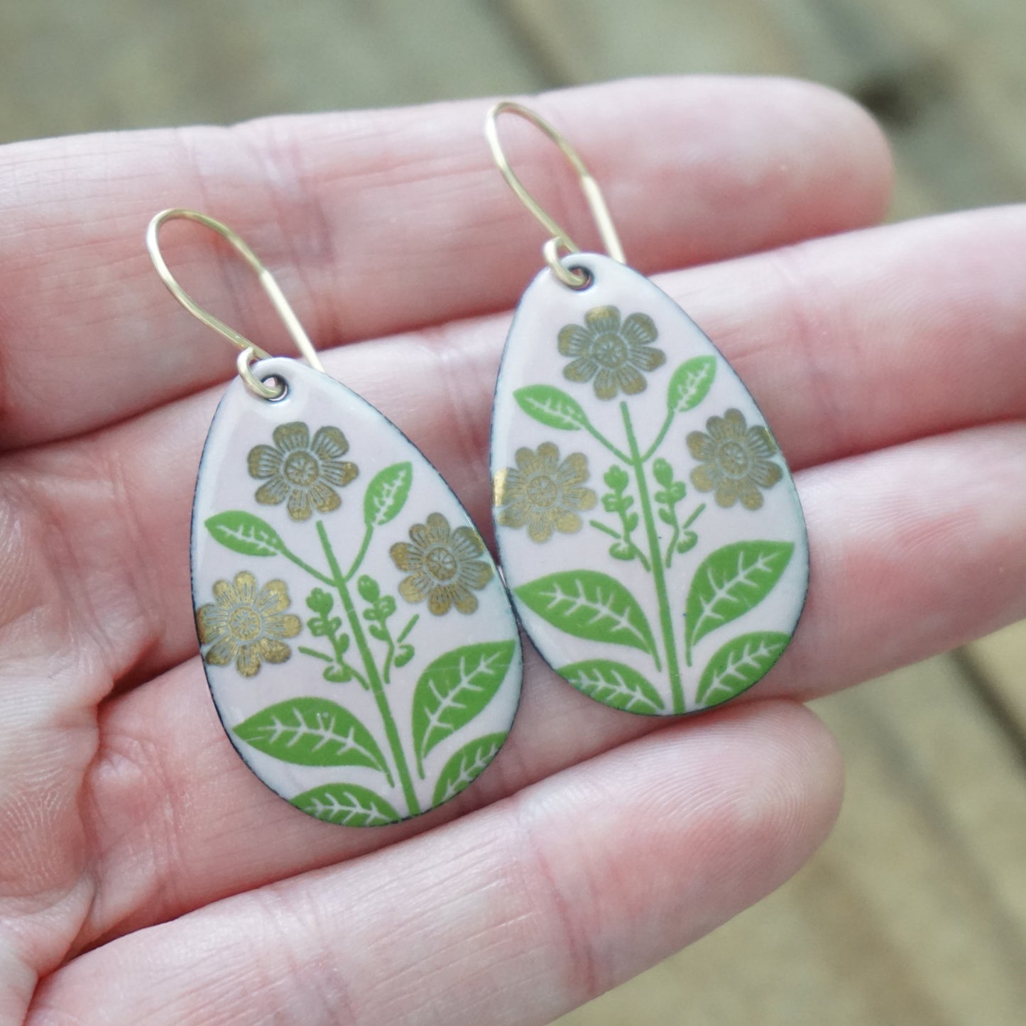 Hand holding a Pink teardrop-shaped dangle earrings, with gold flowers on green stems dangling from gold hooks, against a wooden background
