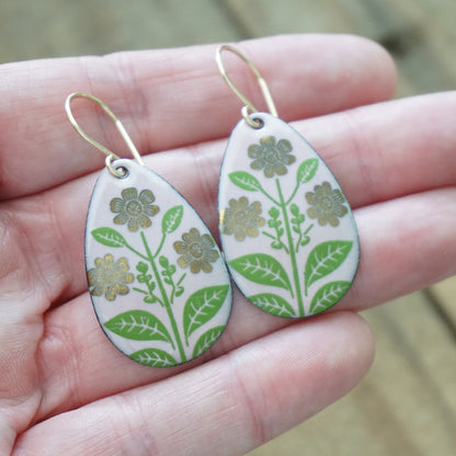 Hand holding a Pink teardrop-shaped dangle earrings, with gold flowers on green stems dangling from gold hooks, against a wooden background