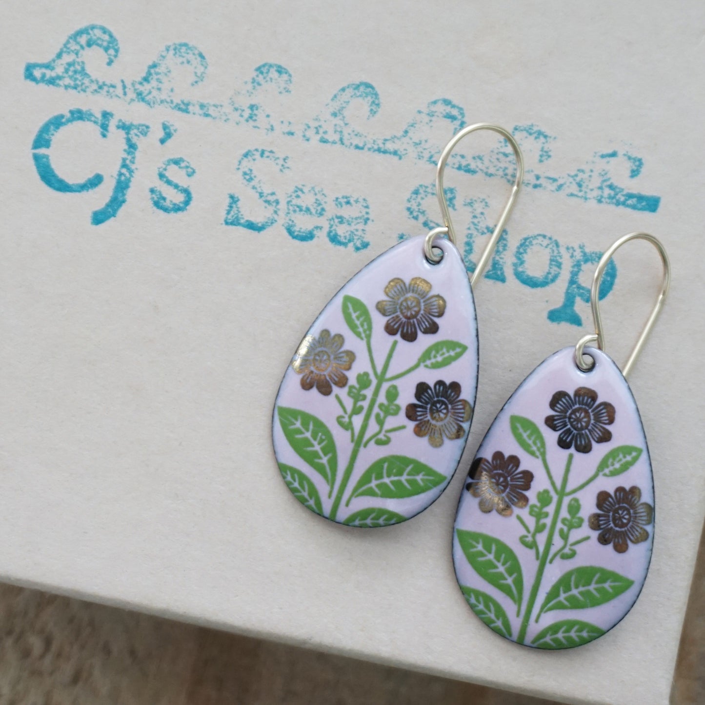 Pink teardrop-shaped dangle earrings, with gold flowers on green stems dangling from gold hooks, displayed on a gift box