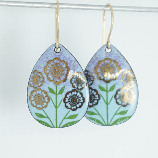 Light blue and purple teardrop-shaped dangle earrings, with gold flowers on green stems dangling from gold hooks, against a white background