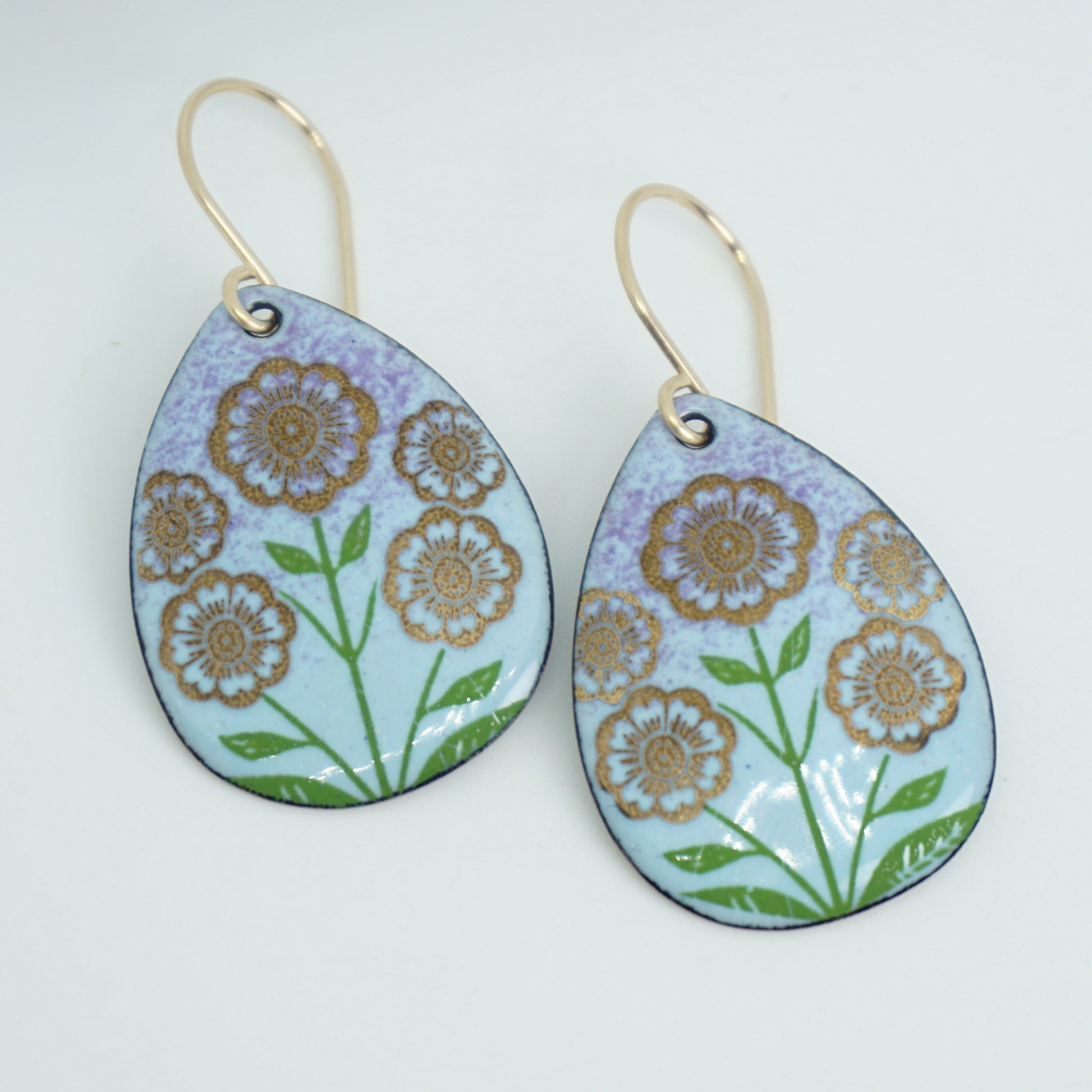 Light blue and purple teardrop-shaped dangle earrings, with gold flowers on green stems dangling from gold hooks, against a white background