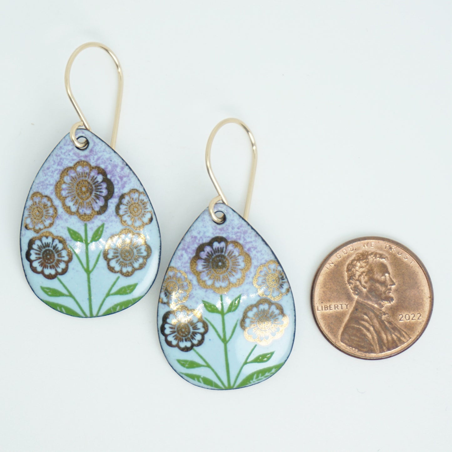 Light blue and purple teardrop-shaped dangle earrings, with gold flowers on green stems dangling from gold hooks, next to a penny for scale, against a white background