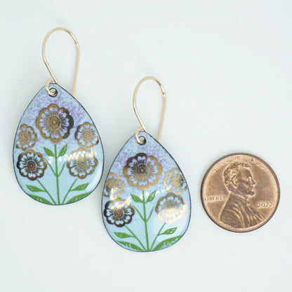 Light blue and purple teardrop-shaped dangle earrings, with gold flowers on green stems dangling from gold hooks, next to a penny for scale, against a white background