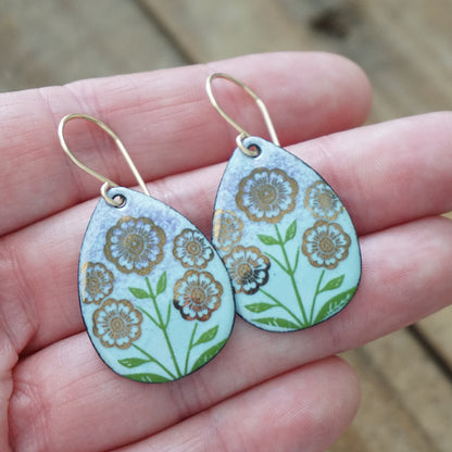 Hand holding a pair of Light blue and purple teardrop-shaped dangle earrings, with gold flowers on green stems dangling from gold hooks, against a wooden background